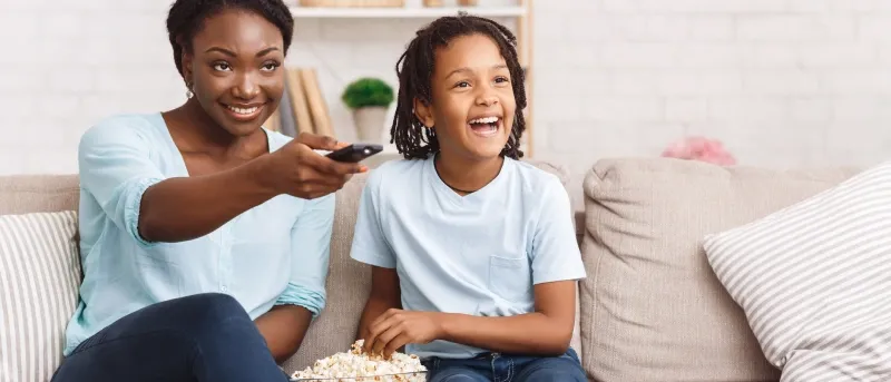 A smiling mother and her son are sitting on a tan sofa, with the mother holding a remote control and the son happily laughing while holding a bowl of popcorn.